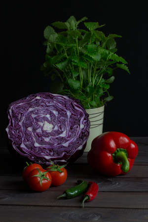 Fresh vegetables red cabbage paprika tomatoes chilli peppermint still life low key. Cut red cabbage, green red hot chili pepper, mint in a glass. Black background, fresh raw vegetables on the tableの写真素材