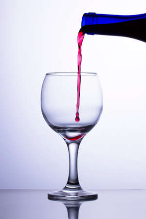 The first drop of wine falls into an empty transparent glass. A jet of red wine flows into a glass from a blue bottle. Wine jet did not reach the glassの写真素材