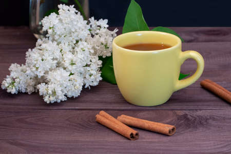 Yellow cup of tea with cinnamon on a dark wooden table decorated with a branch of white lilac. A pleasant tea party with cinnamon sticks and flowers. A cup of fresh Chinese tea.の写真素材