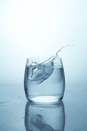 A splash of water from a falling ice cube. A clean glass with drinking water. Drops of water, transparent reflex splash of liquidの写真素材