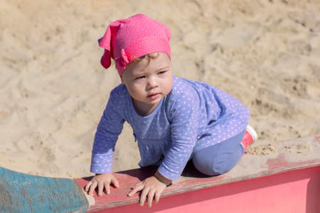 Little girl 1 year old in a blue dress stands on all fours on the edge of the sandbox, summer sunny day walkの写真素材