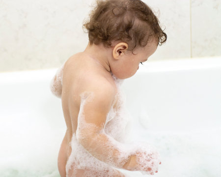 A 1-year-old baby washes in the bath, baby girl boy stands in the water with foam. view from the...