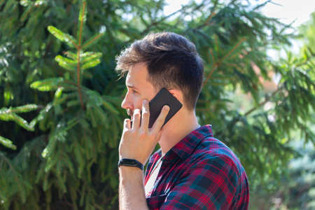Handsome young brunette man is talking on the phone. Profile of caucasian man with a smartphone on the background of spruce evergreen treesの写真素材