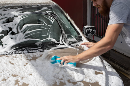 Male motorist washing car with a washcloth and shampoo, auto covered with foam, self-service car...