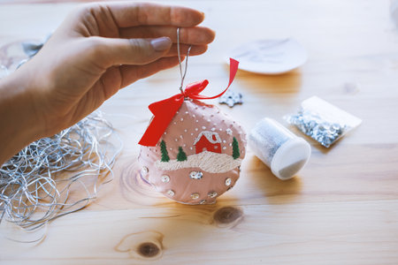 Female hand holding handmade Christmas bauble with needlework and red bow. Close-up Xmas ornament DIY at home. Winter decor from silk and embroidery. Film grain pixel texture. Soft focus.の写真素材
