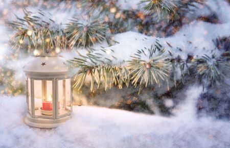Christmas lantern with burning candle shines on snow and spruce branches, snowfall winter scene with light and bokehの写真素材