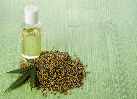 Hemp oil in glass bottle stands on green wooden background with seeds and cannabis leaves, copy spaceの写真素材