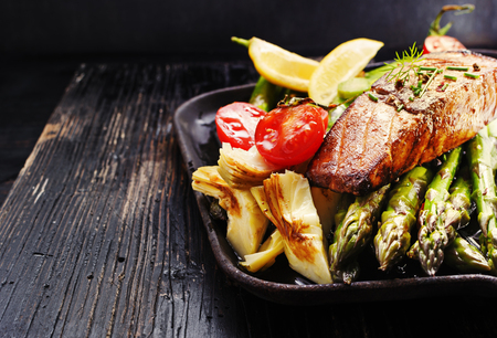 Recipe - delicious portion of grilled salmon fillet with asparagus, tomatoes, artichoke, spices on a black wooden board. Close up, copyspace, selective focus.の写真素材