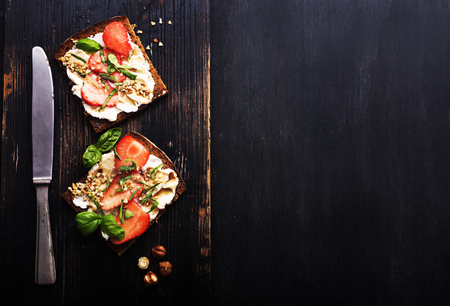 Open sandwiches rye bread with cream cheese and strawberry, basil, hazelnuts, tomatoes on old black metal wooden  surface. Healthy snack concept. Top view.の写真素材