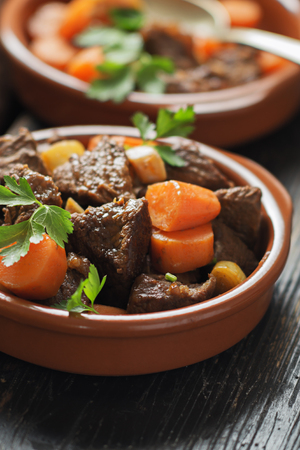 Beef meat stewed with carrots and spices in ceramic pot  on old black wooden board. Selective focus.の写真素材