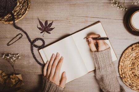 Fall winter background. Female hand holding a pen for a sketch or letter on old notebook with a ball of wool yarn and cup of cappuccino on a wooden background. Home hobbies, Cozy comfort concept.の写真素材