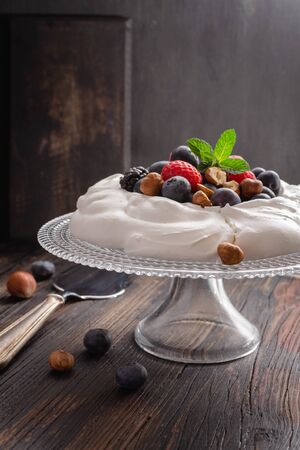 Pavlova cake with fresh summer berries and hazelnuts on glass standcake  on black wooden table.の写真素材