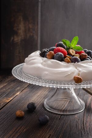 Pavlova cake with fresh summer berries and hazelnuts on glass standcake  on black wooden table.の写真素材