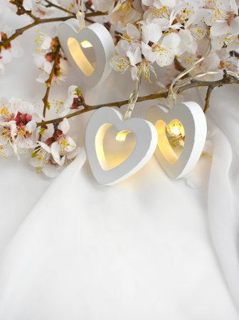 Romantic composition with blooming cherry branch, heart lights on white silk background. Elegant...