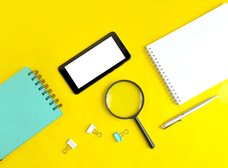 Smartphone, magnifying glass, notebook and stationery on yellow background. Job or information...