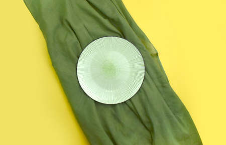 Empty ceramic plate with green fabric on yellow background. Minimalist summer table setting. Eco friendly style. Trendy color design. Lined ornament. Top view.の写真素材