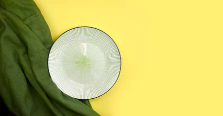 Empty ceramic plate with green fabric on yellow background. Minimalist summer table setting. Eco friendly style. Trendy color design. Top view. Bannerの写真素材