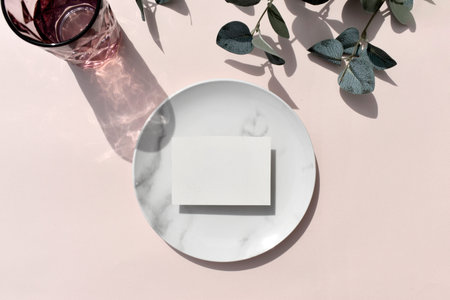 Mockup empty blank invitation card, marble plate color glass and with eucalyptus leaves on sunlit beige background. Elegant minimal tableware composition for wedding, restaurant menu or business.の写真素材