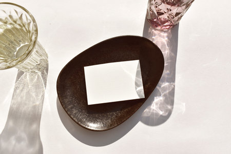 Luxury summer mockup table setting. Empty paper blank on stoneware plate, cocktail glass on sunlit background. Mock invitation card for wedding, business or menu. Eco minimal wabi sabi styleの写真素材