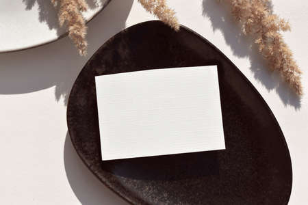 Autumn mockup still life. Empty blank greeting card on black tray with harsh shadows on neutral beige background. Fall minimal table setting for wedding invitation, business or menu.の写真素材