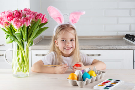 Cute smiling girl with bunny ears paints Easter eggs at the table.の写真素材