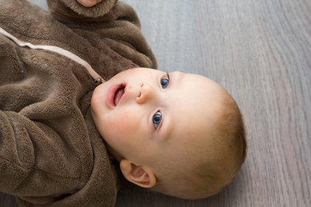 Adorable 9 month boy in brown fur hoody is laying on the grey floor