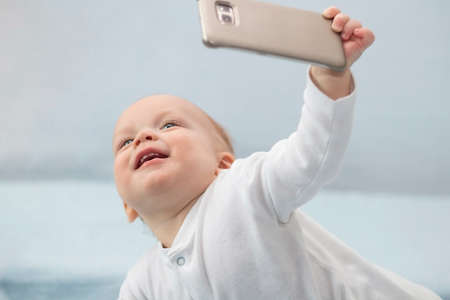 Cute infant boy makes selfie with a cell phone. Adorable smiling toddler kid taking a selfie photo with smartphone.の写真素材
