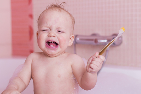 Crying baby boy with a teethbrush in a bathtub.の写真素材