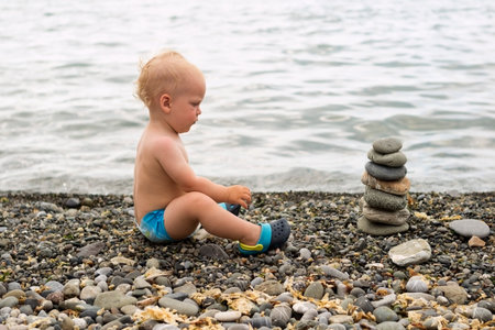 Tranquil toddler meditating on the sea beach. Cute baby boy relaxing and looking to the stone...