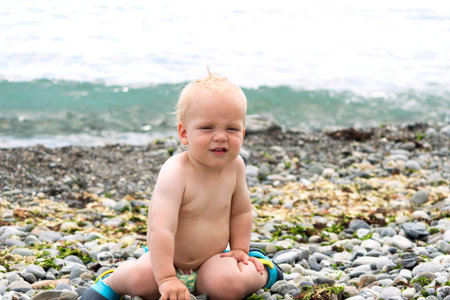 Cute by boy sitting on the stony beach. Funny toddler on the seashore looking at cameraの写真素材
