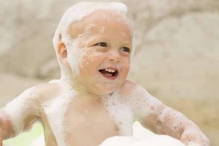 Funny laughing toddler in a soap foam taking a bath against concrete wallの写真素材