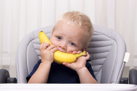 Cute one year old toddler eating a banana and sitting in the baby chair. Funny kid with banana smileの写真素材