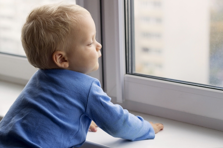 Adorable little blond kid boy sitting near window and looking outdoorsの写真素材