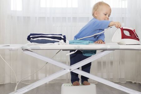 Funny cute little baby boy housekeeper ironing clothes. Kid engaged in domestic workの写真素材