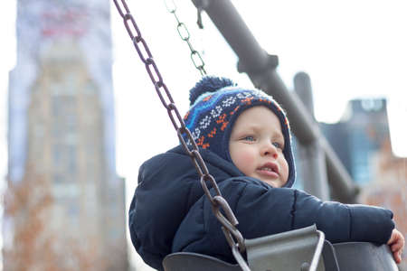 Cute toddler on a swing on the playground against NY skyscrapers. Spring or autumn shot.の写真素材