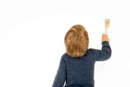 Baby boy with paint brush standing back near white wall