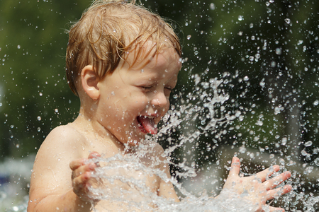 Funny baby boy trying to cauch water stream in fountain. Cute toddler playing in the city fountainの写真素材