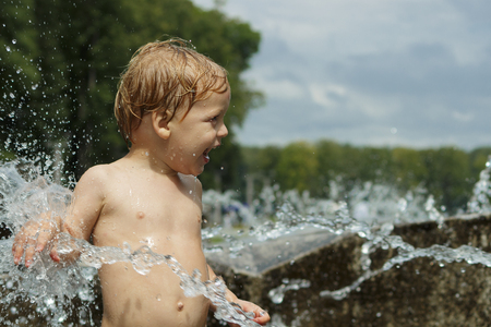 Funny baby boy trying to cauch water stream in fountain. Cute toddler playing in the city fountainの写真素材