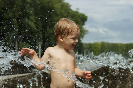 Funny baby boy trying to cauch water stream in fountain. Cute toddler playing in the city fountainの写真素材