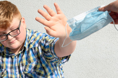The red-haired boy with glasses refuses to receive the surgical mask offered by an adult. Boy with glasses and plaid shirt who refuses to wear a surgical mask.の写真素材