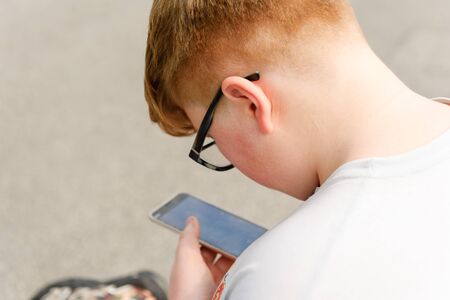 View from behind of a red-haired boy with cellphone and glasses in an urban context. Boy wearing glasses holds smartphone in hand.の写真素材