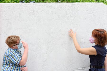 Young red-haired woman and boy wearing a surgical mask stand against a background of the wall with the view of green trees.の写真素材