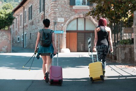 Hand suitcase. Two women walking and rolling her wheeled suitcases on the sidewalk. Luggage is located on the city street and is ready for travel. Rear view of two girls traveling.の写真素材