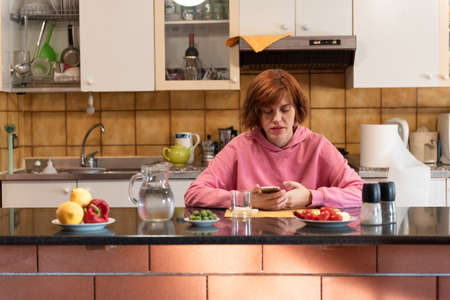 Young cute woman eats in her kitchen and holding smartphone.の写真素材