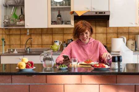 Young cute woman eats in her kitchen and holding mobile phone.の写真素材