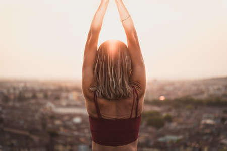 Front view of a back light of a faithful woman in yoga position holding the sun at sunset or sunrise in a beautiful city location with a warm backgroundの写真素材