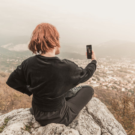 The red-haired girl taking photo with smart phone at the mountain peak