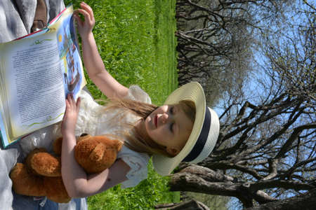 little girl reading a book in the park with her teddy bearの写真素材