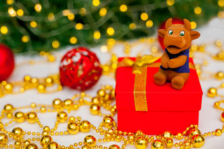 Toy brown bull on a red gift box on a white background. Christmas tree branch with red toys and golden garlands in the background. Bull as a symbol of the new year 2021. New Year, Christmas conceptの写真素材
