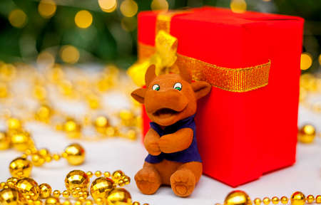 Toy brown bull on a red gift box on a white background. Christmas tree branch with red toys and golden garlands in the background. Bull as a symbol of the new year 2021. New Year, Christmas conceptの写真素材